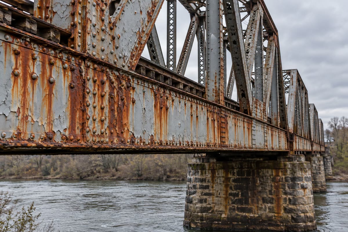 Uma velha ponte ferroviária rebitada. Escorrimentos de ferrugem descem dos rebites e das juntas, onde a água e o oxigênio permanecem por mais tempo e a pintura falha primeiro.