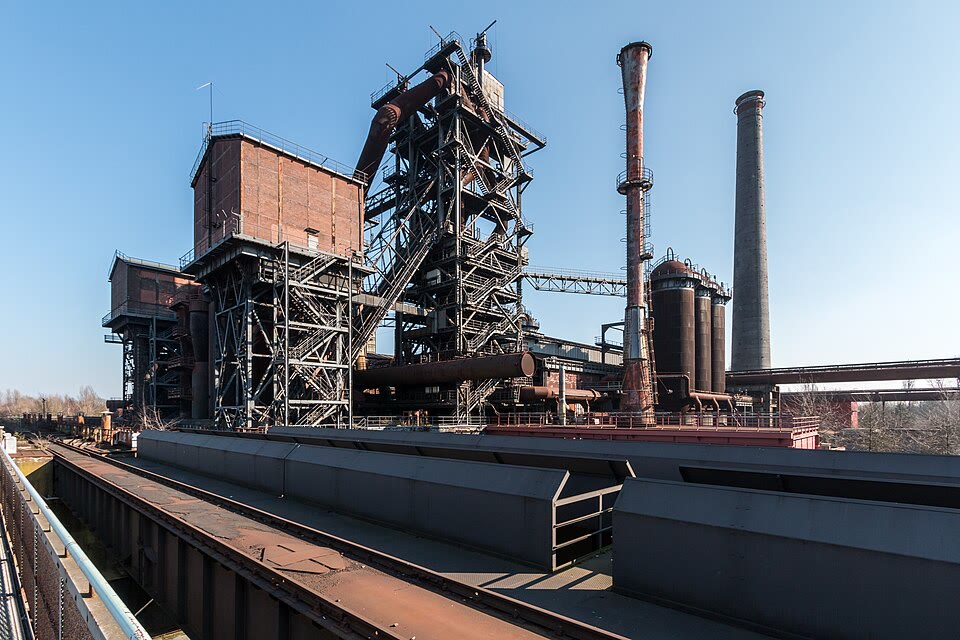 A blast furnace preserved as a monument (Duisburg-Nord, shut down in 1985): the tall shaft with its ring of pipes for the hot air, the gas off-takes at the top. (Photograph: Dietmar Rabich, CC BY-SA 4.0, Wikimedia Commons.)