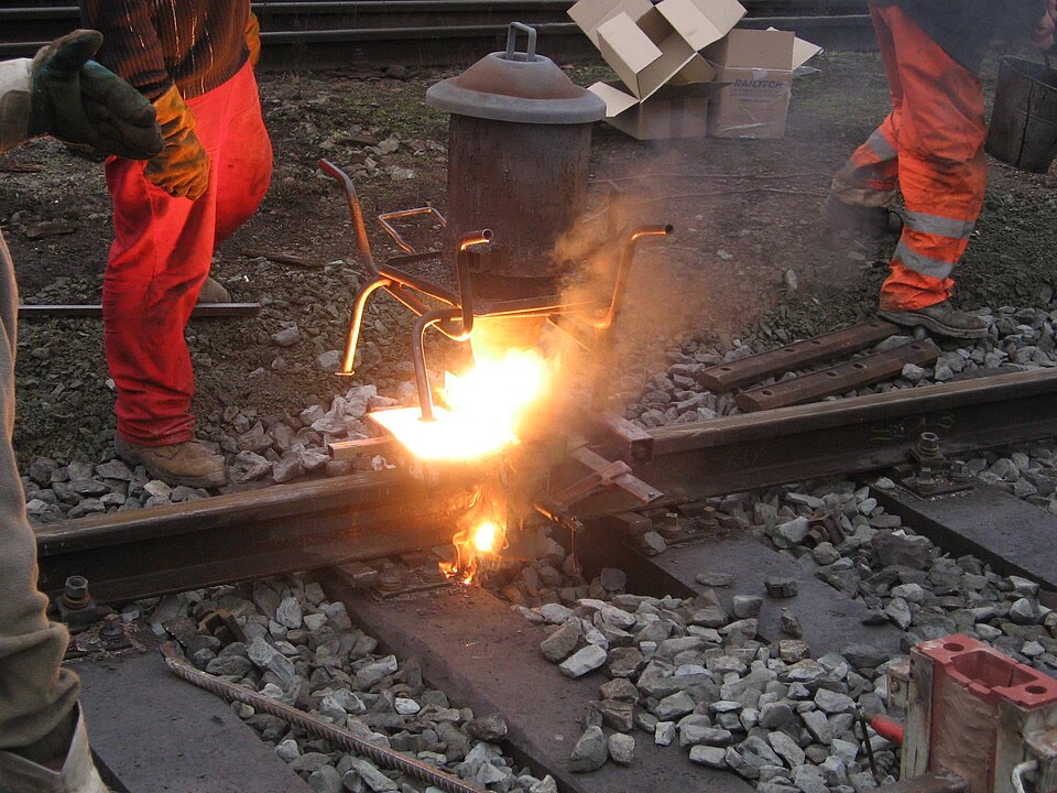 Thermite welding of a rail: the reaction of iron oxide with aluminium in the crucible above the joint gives liquid iron, which runs down into the mould between the two rail ends. (Photograph: PetrS., CC BY-SA 3.0, Wikimedia Commons.)